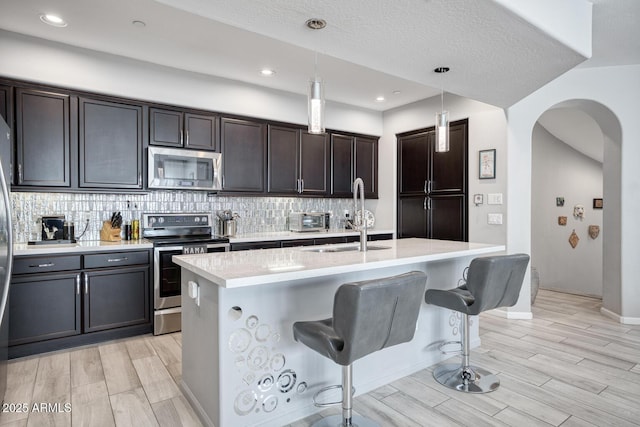 kitchen featuring a kitchen island with sink, appliances with stainless steel finishes, a sink, and decorative light fixtures