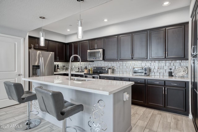 kitchen with decorative light fixtures, appliances with stainless steel finishes, a kitchen island with sink, a sink, and dark brown cabinets