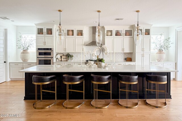 kitchen with wall chimney exhaust hood, double oven, light countertops, tasteful backsplash, and a large island with sink