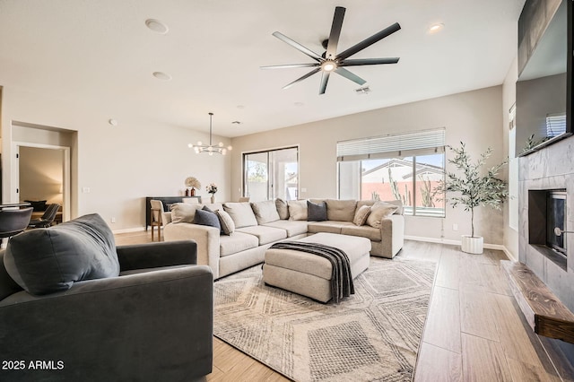 living room with light wood-type flooring, baseboards, a tiled fireplace, and ceiling fan with notable chandelier