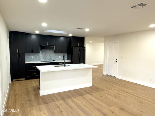 kitchen with sink, decorative backsplash, black appliances, a center island with sink, and light hardwood / wood-style flooring