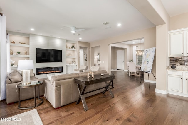 living room with dark wood-type flooring, a large fireplace, ceiling fan, and built in shelves