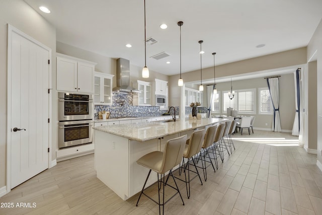 kitchen featuring appliances with stainless steel finishes, white cabinetry, hanging light fixtures, wall chimney range hood, and a spacious island