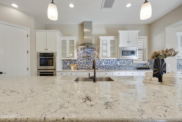 kitchen featuring wall chimney range hood, appliances with stainless steel finishes, hanging light fixtures, tasteful backsplash, and light stone countertops