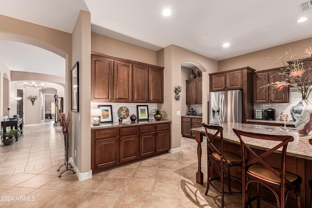 kitchen with light stone counters, backsplash, dark brown cabinetry, a kitchen bar, and stainless steel refrigerator with ice dispenser