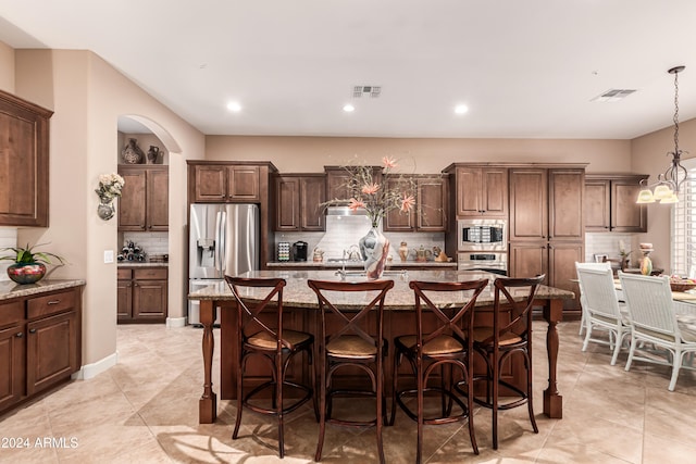 kitchen featuring stainless steel appliances, an island with sink, backsplash, light stone countertops, and pendant lighting