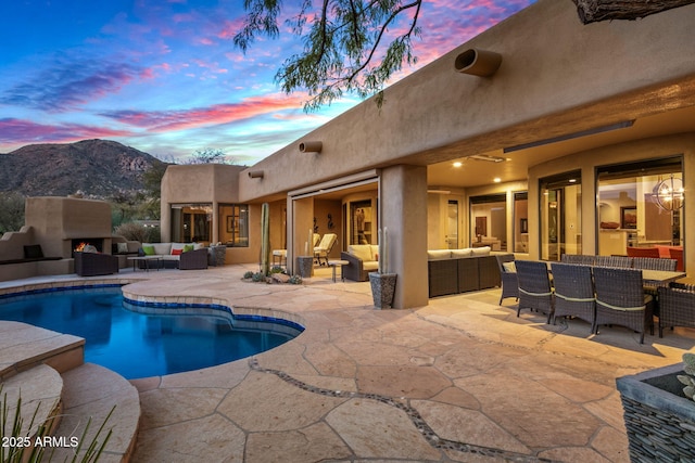 pool at dusk with an outdoor living space with a fireplace, a mountain view, and a patio area