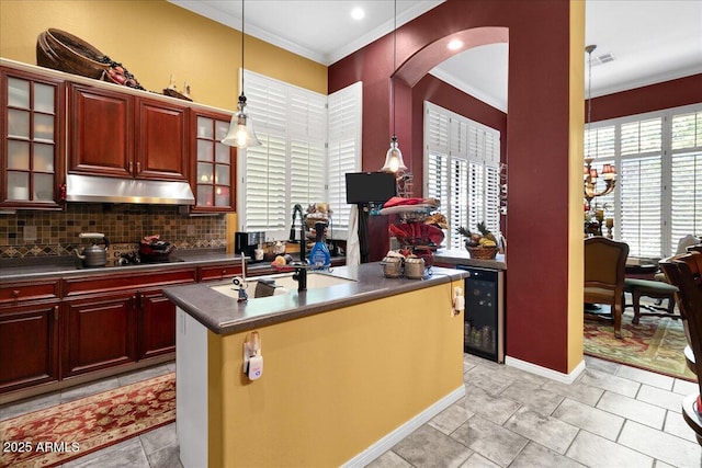 kitchen with under cabinet range hood, dark countertops, ornamental molding, dark brown cabinets, and beverage cooler