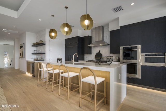 kitchen with an island with sink, wall chimney exhaust hood, light wood-type flooring, and pendant lighting