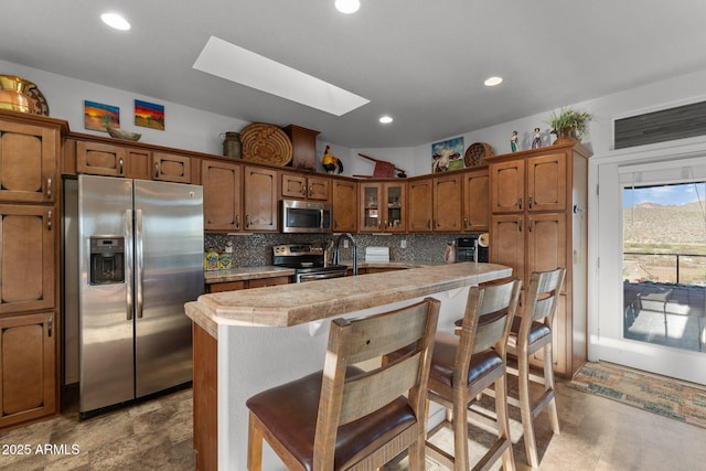 3874 Pura Vida Trl, Wickenburg, AZ 85390 - kitchen with stainless steel appliances, a breakfast bar, backsplash, a skylight, and brown cabinetry