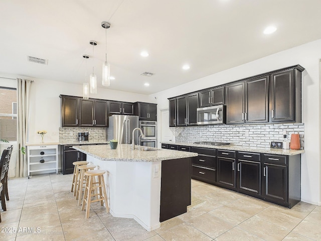 kitchen featuring light tile patterned flooring, appliances with stainless steel finishes, hanging light fixtures, a kitchen island with sink, and light stone countertops