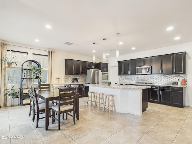 kitchen with sink, appliances with stainless steel finishes, light stone countertops, a center island with sink, and decorative light fixtures