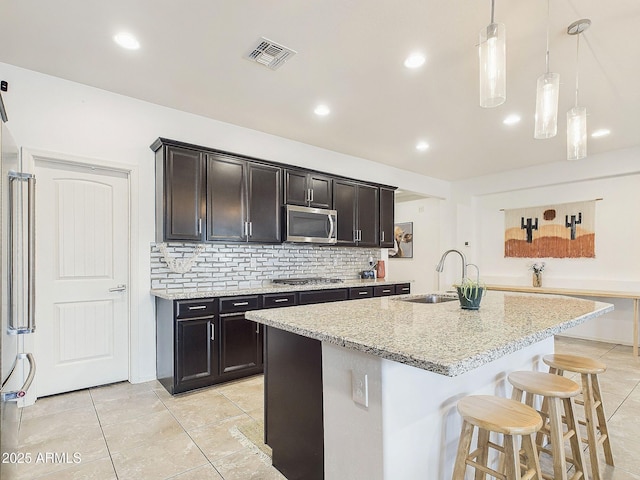 kitchen with sink, a center island with sink, appliances with stainless steel finishes, pendant lighting, and light stone countertops