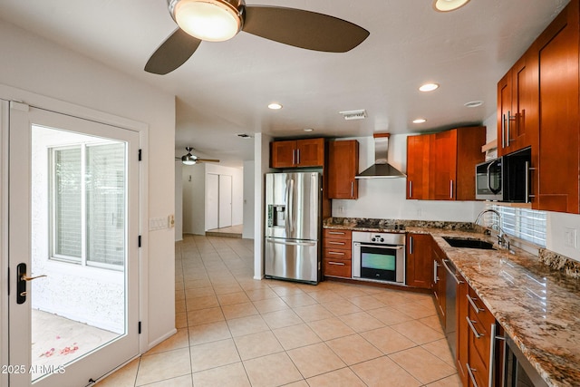 kitchen with a sink, stainless steel appliances, wall chimney range hood, stone counters, and ceiling fan