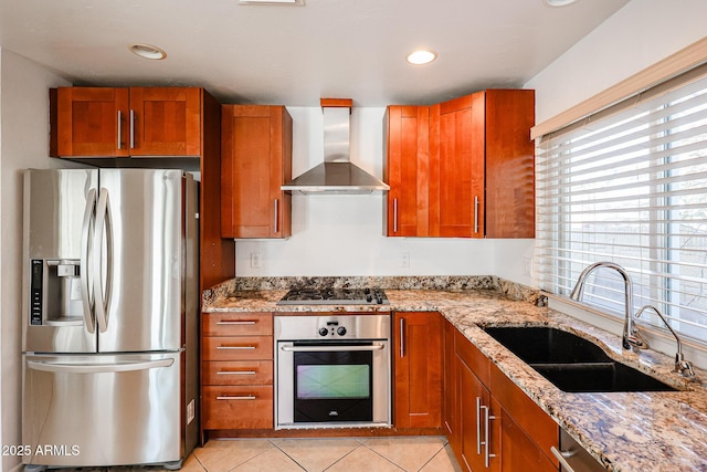 kitchen featuring light stone counters, brown cabinets, appliances with stainless steel finishes, wall chimney exhaust hood, and a sink