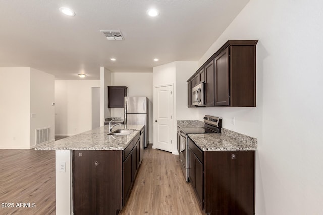 kitchen with an island with sink, sink, light stone counters, stainless steel appliances, and light wood-type flooring