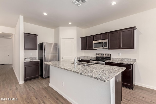 kitchen featuring sink, stainless steel appliances, light stone countertops, an island with sink, and light wood-type flooring