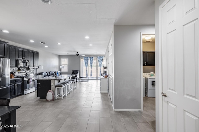 kitchen with dark cabinets, appliances with stainless steel finishes, washing machine and dryer, a breakfast bar, and a ceiling fan