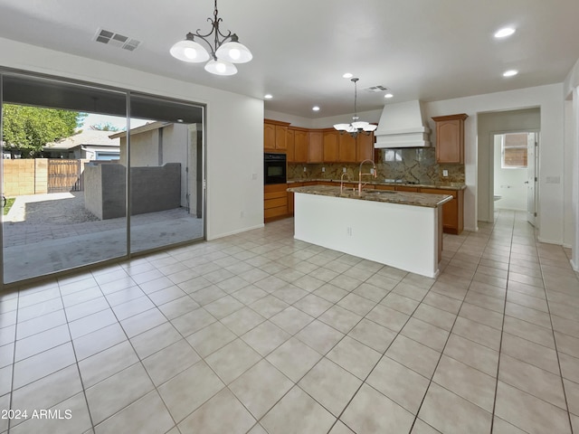 kitchen with custom exhaust hood, hanging light fixtures, a kitchen island with sink, and oven