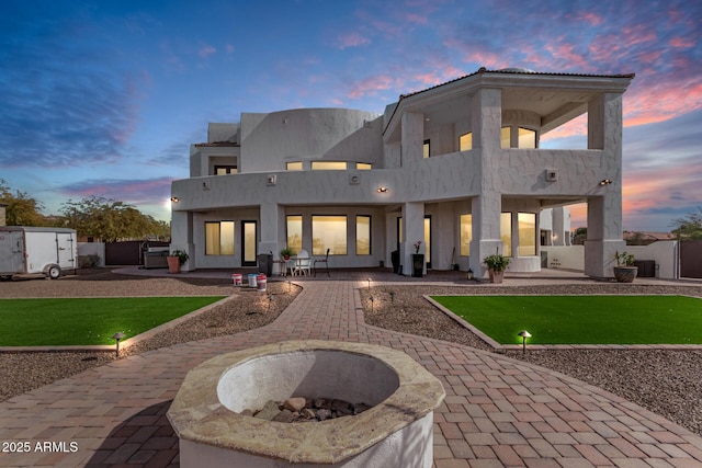 back of house at dusk featuring a balcony, a patio area, an outdoor fire pit, and stucco siding