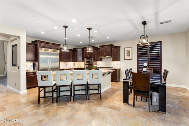 kitchen with dark brown cabinets, appliances with stainless steel finishes, an island with sink, pendant lighting, and backsplash