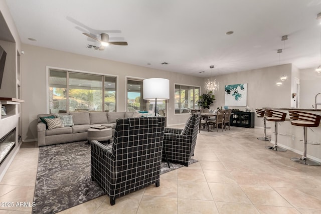 living area with light tile patterned flooring, a glass covered fireplace, ceiling fan with notable chandelier, baseboards, and visible vents
