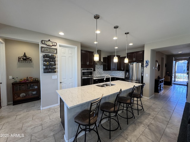 kitchen featuring wall chimney exhaust hood, an island with sink, light stone counters, pendant lighting, and sink