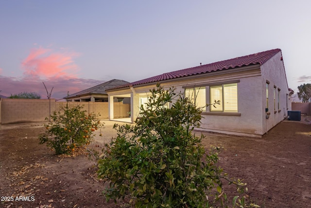 property exterior at dusk featuring a tiled roof, central AC unit, stucco siding, a fenced backyard, and a patio