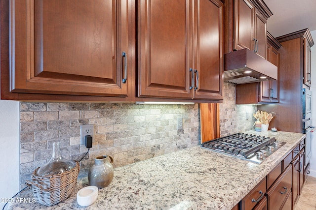 13719 W Amberwing St, Peoria, AZ 85383 - kitchen featuring stainless steel gas cooktop, custom range hood, light stone countertops, decorative backsplash, and brown cabinetry