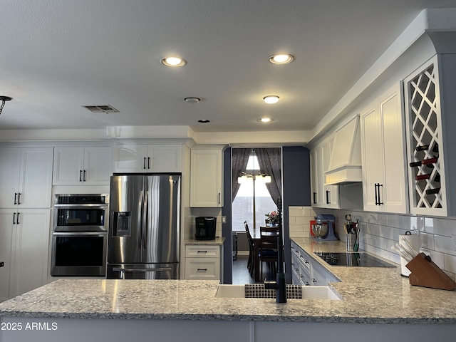 kitchen with tasteful backsplash, white cabinets, custom range hood, light stone countertops, and stainless steel appliances