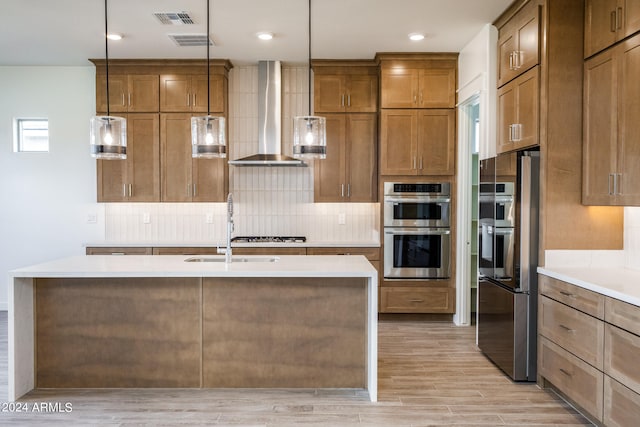 kitchen with light wood-type flooring, wall chimney exhaust hood, stainless steel double oven, decorative light fixtures, and an island with sink