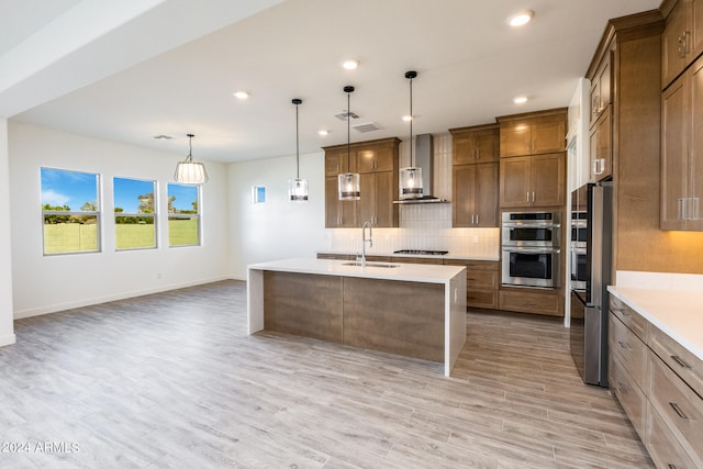 kitchen with light wood-type flooring, gas stovetop, wall chimney range hood, hanging light fixtures, and an island with sink