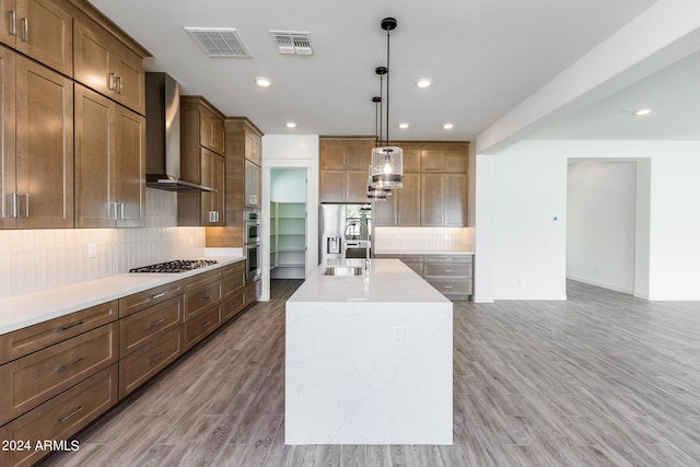 kitchen with wall chimney exhaust hood, light hardwood / wood-style floors, an island with sink, and appliances with stainless steel finishes