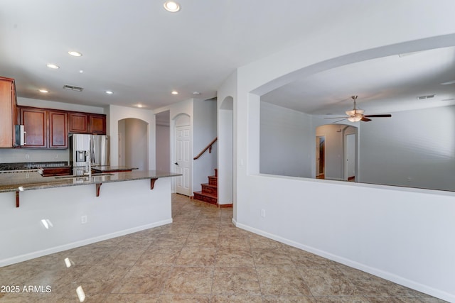 kitchen featuring dark stone counters, arched walkways, ceiling fan, a kitchen bar, and stainless steel fridge