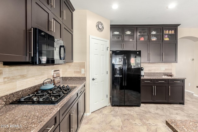 kitchen with tasteful backsplash, arched walkways, glass insert cabinets, black appliances, and stone counters