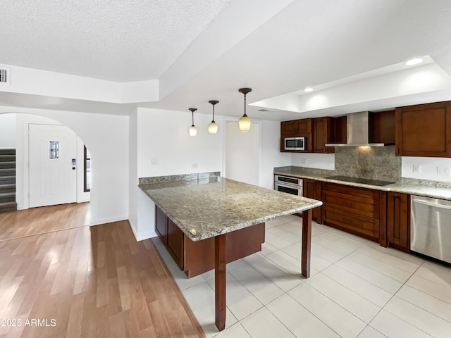 kitchen featuring tasteful backsplash, a raised ceiling, arched walkways, stainless steel appliances, and wall chimney exhaust hood