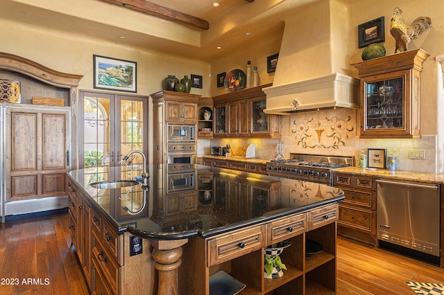 kitchen featuring appliances with stainless steel finishes, premium range hood, sink, a center island with sink, and hardwood / wood-style floors