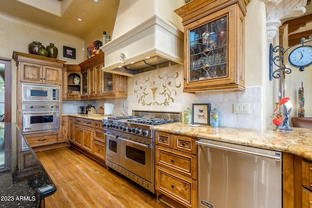 kitchen with light stone countertops, stainless steel appliances, custom range hood, and light wood-type flooring