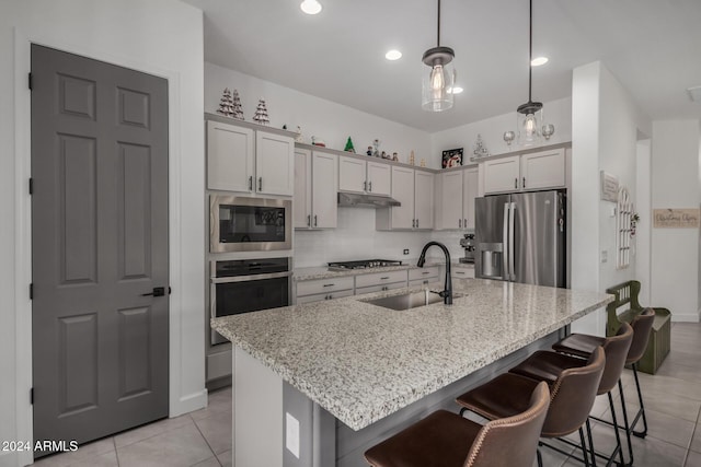 kitchen with a kitchen island with sink, hanging light fixtures, sink, light stone countertops, and stainless steel appliances