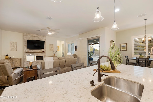 kitchen with light stone countertops, pendant lighting, sink, a fireplace, and ceiling fan with notable chandelier