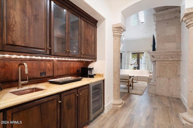 bar with dark brown cabinets, beverage cooler, decorative columns, light hardwood / wood-style flooring, and sink