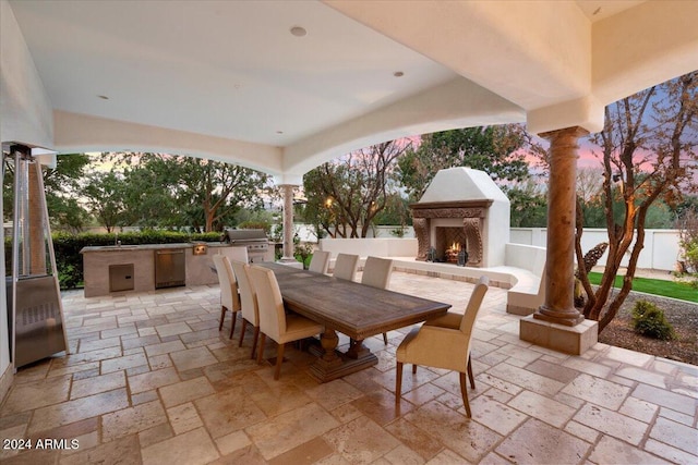 patio terrace at dusk with exterior fireplace, an outdoor kitchen, and a grill