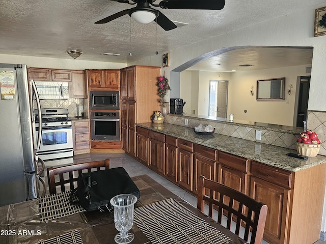 kitchen featuring a textured ceiling, appliances with stainless steel finishes, kitchen peninsula, light stone countertops, and backsplash