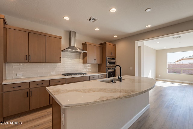 kitchen featuring backsplash, wall chimney range hood, light hardwood / wood-style flooring, an island with sink, and stainless steel appliances