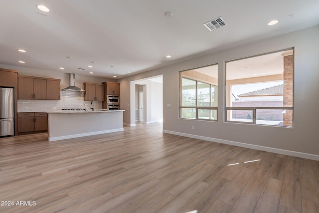 kitchen with stainless steel appliances, wall chimney exhaust hood, an island with sink, and light wood-type flooring