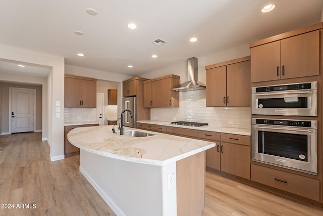 kitchen featuring appliances with stainless steel finishes, light wood-type flooring, wall chimney exhaust hood, a kitchen island with sink, and sink