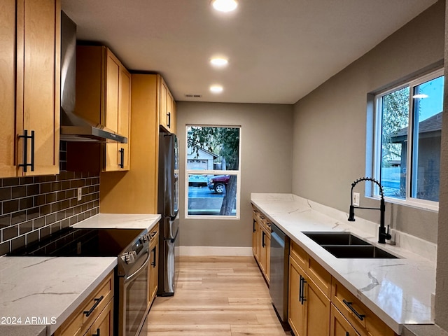 kitchen with appliances with stainless steel finishes, light stone counters, wall chimney exhaust hood, light hardwood / wood-style flooring, and sink