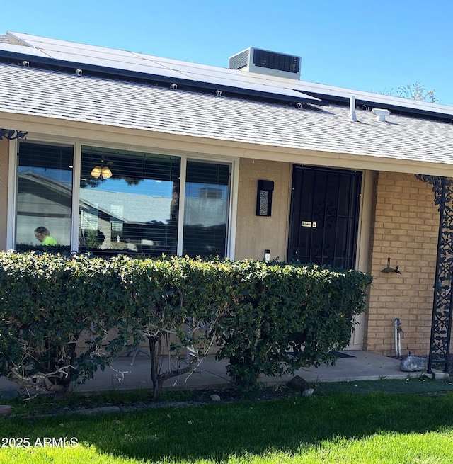 view of exterior entry with brick siding, stucco siding, a shingled roof, central AC unit, and roof mounted solar panels