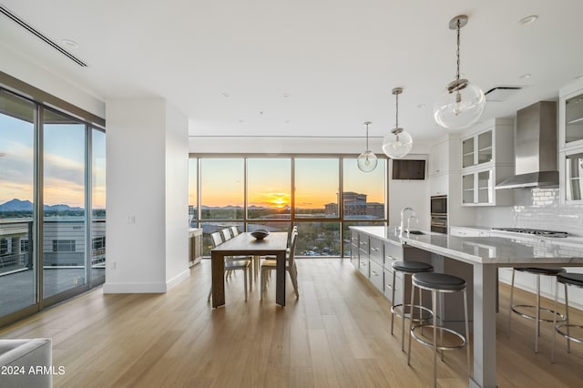 kitchen with wall chimney range hood, light wood-type flooring, tasteful backsplash, decorative light fixtures, and a kitchen bar