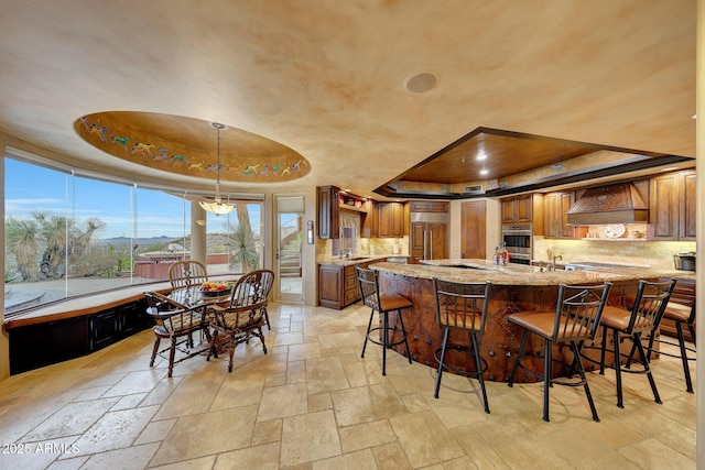 kitchen featuring appliances with stainless steel finishes, a breakfast bar, a raised ceiling, light stone countertops, and custom range hood